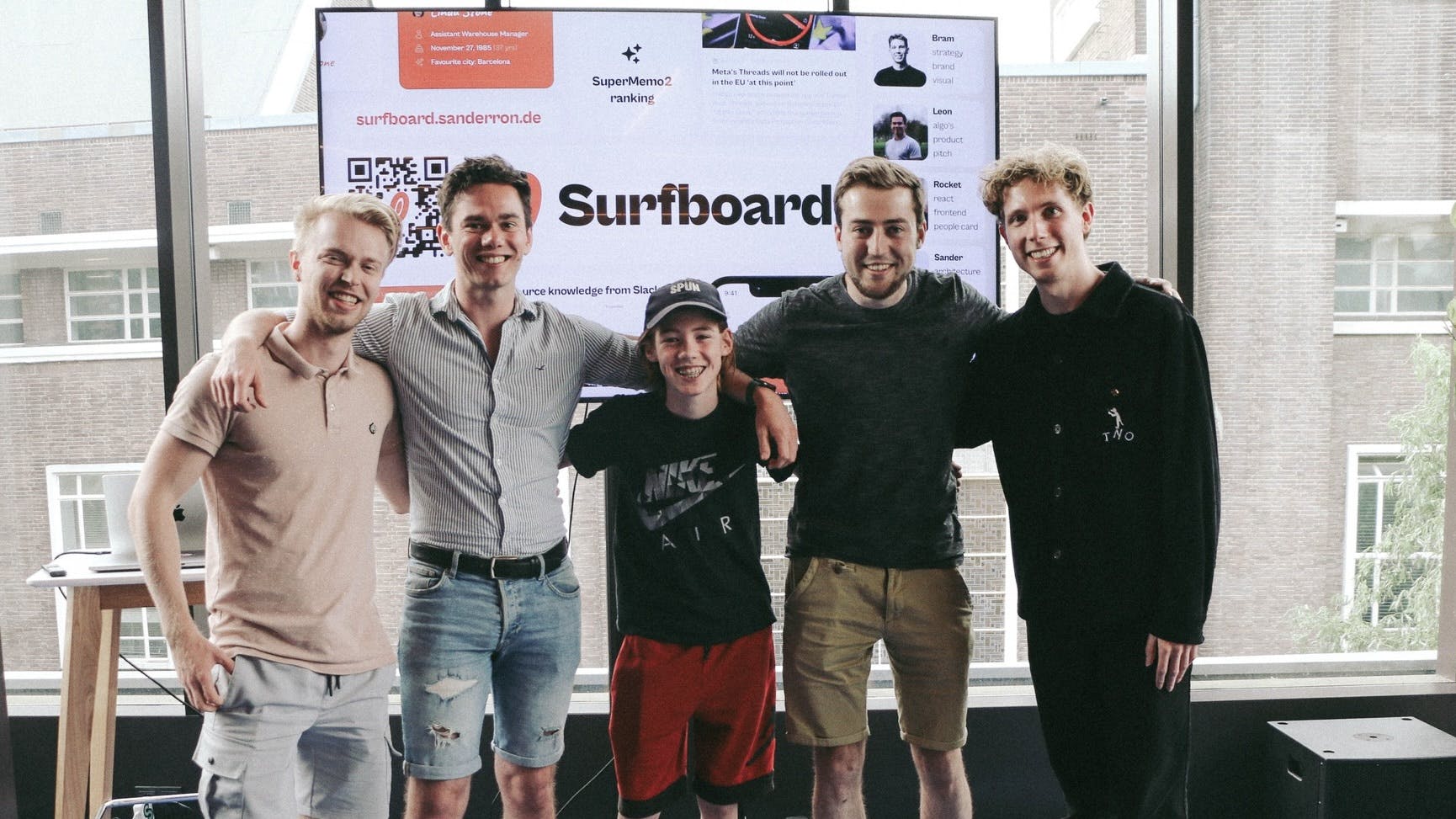 A picture of the five team members standing in front of a big television screen showing a slide with the Surfboard logo on it. They are holding each other’s shoulders and smiling at the camera. From left to right are myself, Leon, Rocket, Sander and Bram.