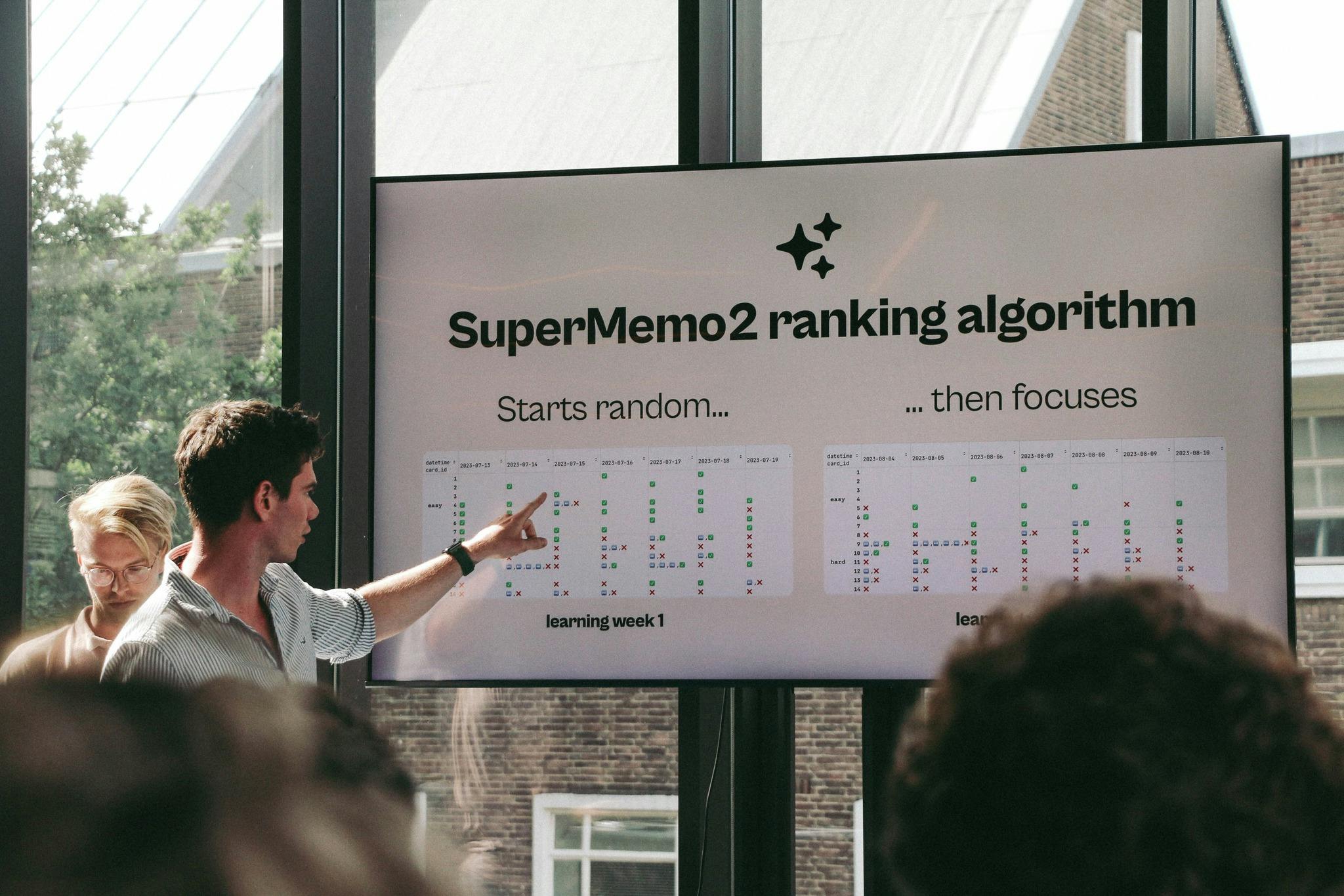 A picture of Leon pointing to a big television screen. On the screen, our pitch deck is showing a slide titled “SuperMemo2 ranking algorithm” with two columns of data: “Starts random…” and “…then focuses”.